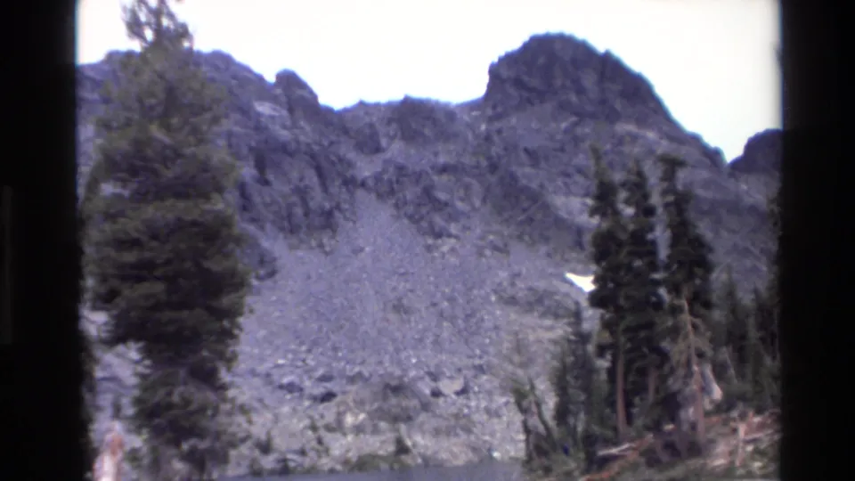 Talus-covered mountain slope with tall conifers and jagged peaks, California mountain area circa 1980, 8mm home movie