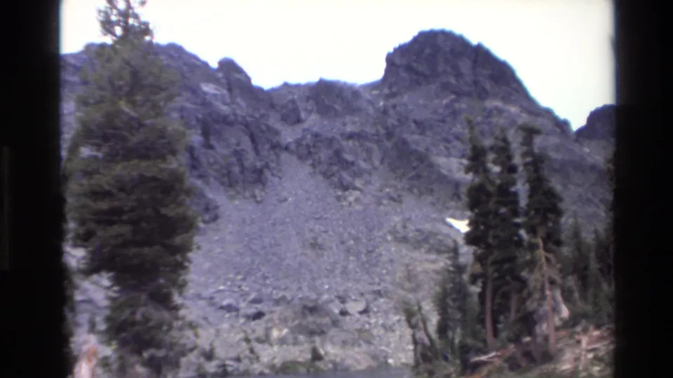 Talus slope beneath dark jagged peaks with conifers and residual snow patch, California mountain wilderness circa 1980, scanned 8mm