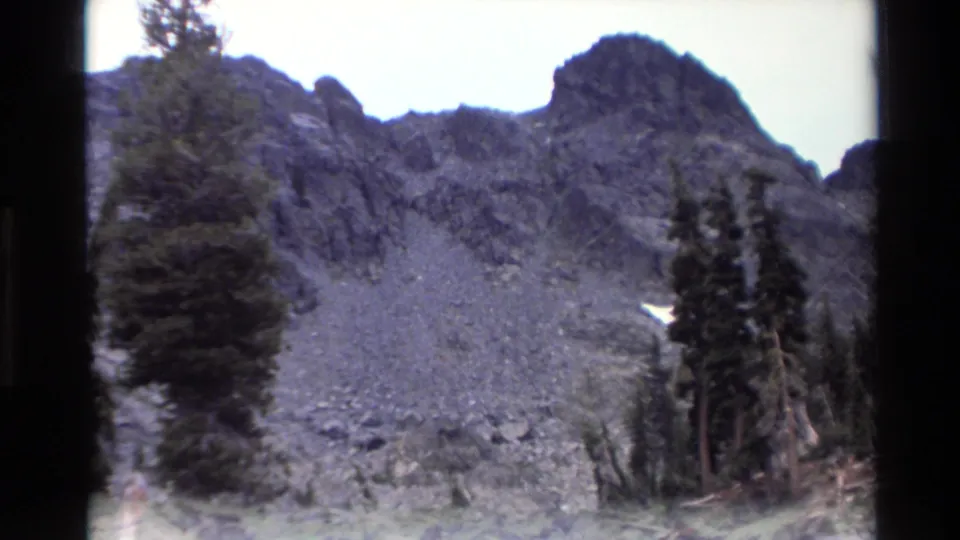 Pan continues across rocky talus slope with conifers and snow remnant, California mountain area circa 1980, 8mm home movie