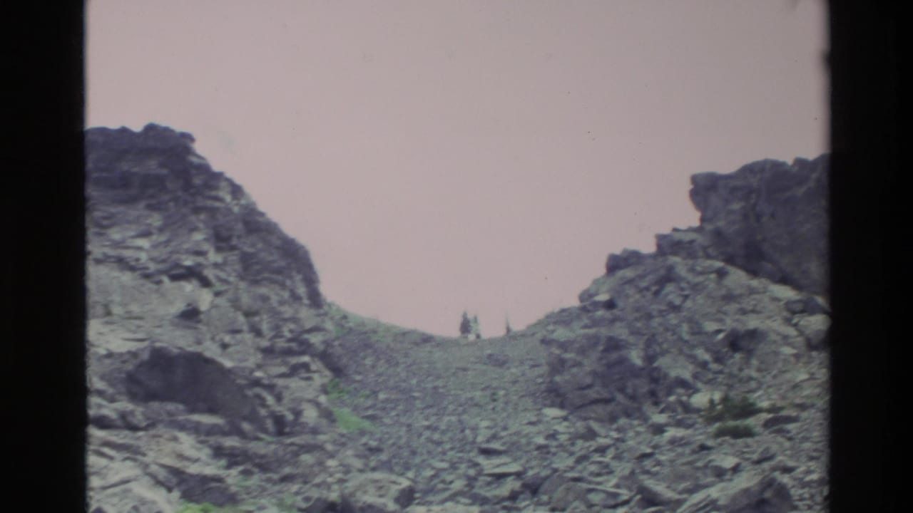 Super 8 home movie frame showing a rugged scree slope at Big Sur, California, circa 1980, with two small figures visible near a rocky ridge saddle.