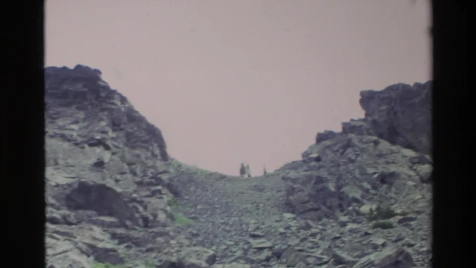 Super 8 home movie frame showing a rugged scree slope at Big Sur, California, circa 1980, with two small figures visible near a rocky ridge saddle.