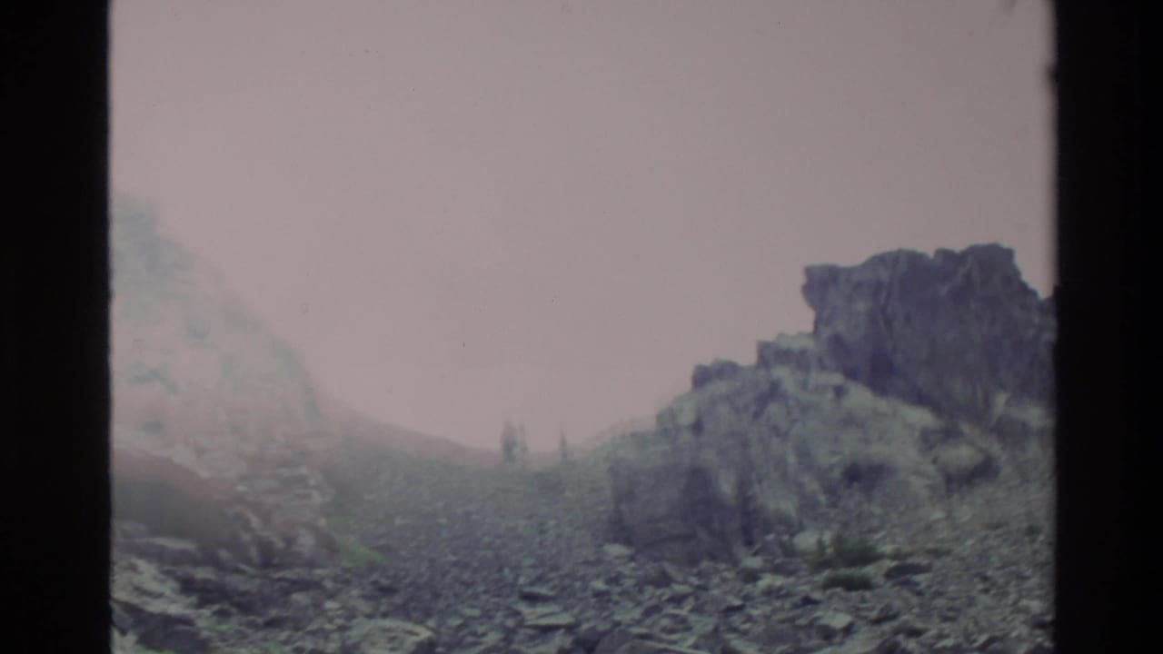 Rocky mountain slope with fog or haze in upper left, dark outcropping on right, pale overcast sky.