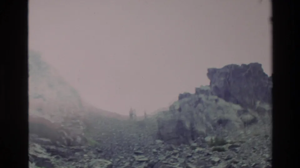 Super 8 frame of a scree and rock slope, likely Big Sur, California circa 1980, with misty sky and dark jagged rock formations.