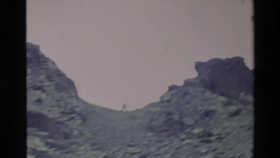 Super 8 home movie frame of rugged dark rock formations and loose scree, Big Sur area, California, circa 1980, under a pale overcast sky.