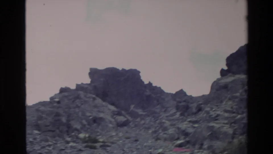 Super 8 frame showing a dark rocky ridge against a pale pink overcast sky, Big Sur area, California, circa 1980, with a film edge artifact at lower right.