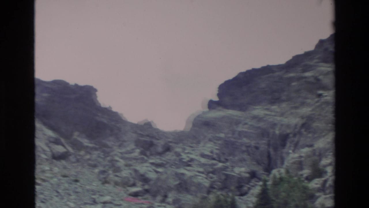 Close-up pan of a steep rocky mountain face with conifers at its base, Big Sur area, California, c. 1980, with pinkish Super 8 color cast.