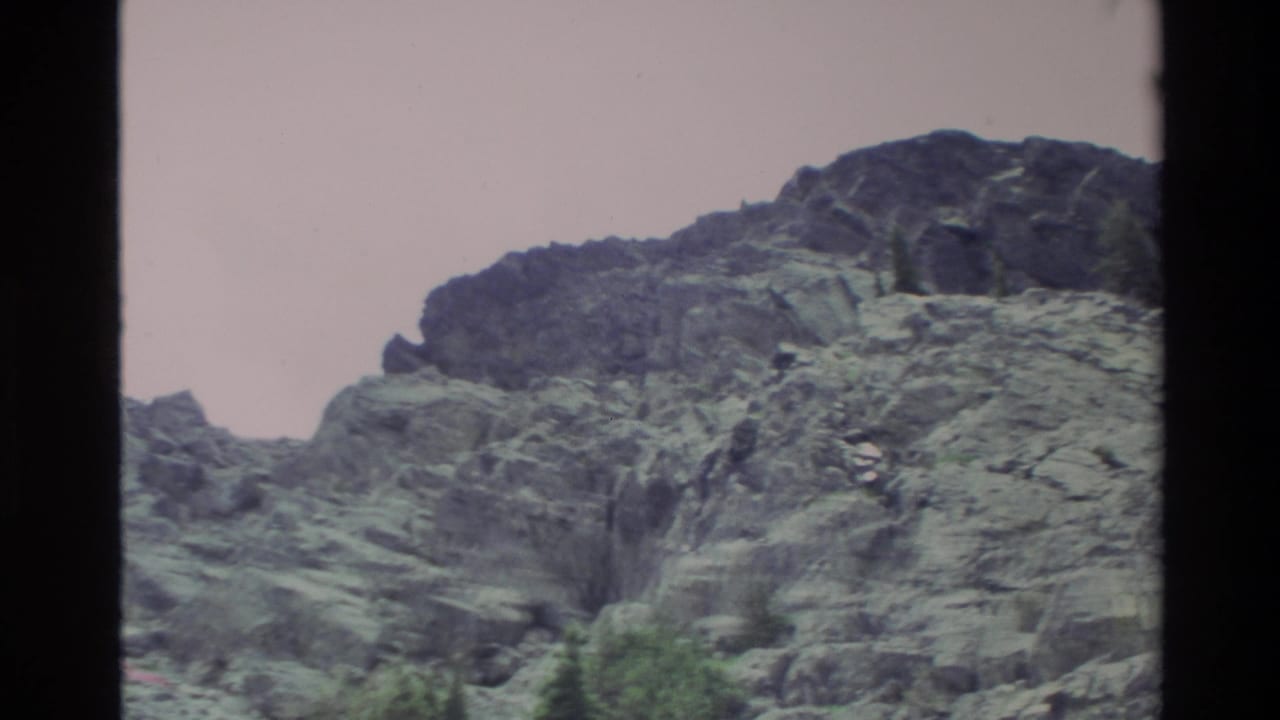Rocky mountain cliff face mid-pan, Big Sur area, California, c. 1980; conifers at lower frame edge, pinkish Super 8 color cast.