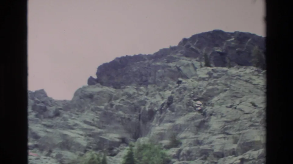 Rocky mountain cliff face mid-pan, Big Sur area, California, c. 1980; conifers at lower frame edge, pinkish Super 8 color cast.