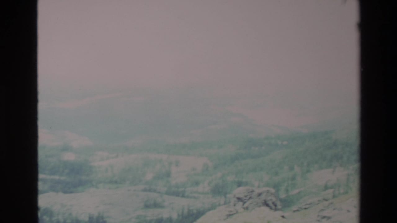 Early frame of a slow pan across Big Sur-area hills in apparent 1980s 8mm home movie; overcast sky, rocky foreground, forested valley below.