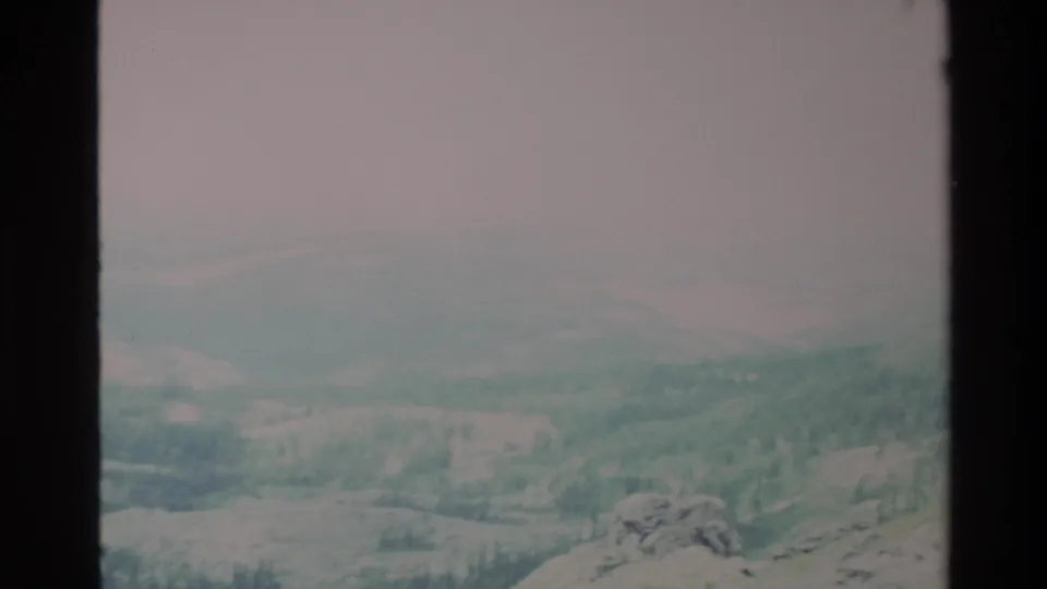 Early frame of a slow pan across Big Sur-area hills in apparent 1980s 8mm home movie; overcast sky, rocky foreground, forested valley below.