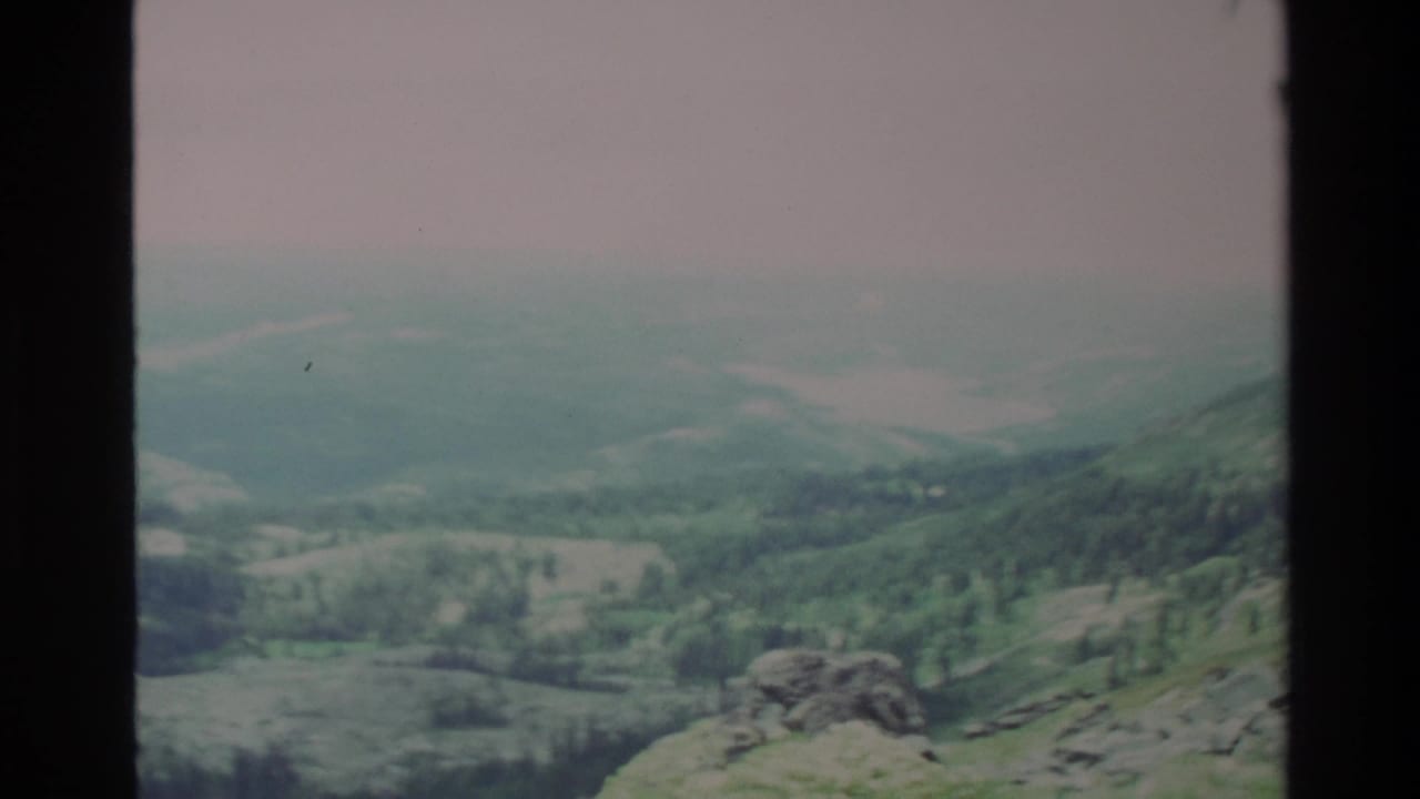 Mid-pan frame from apparent 1980s 8mm footage of Big Sur area; rocky outcrop and dense conifers in mid-ground, pale hazy sky above.