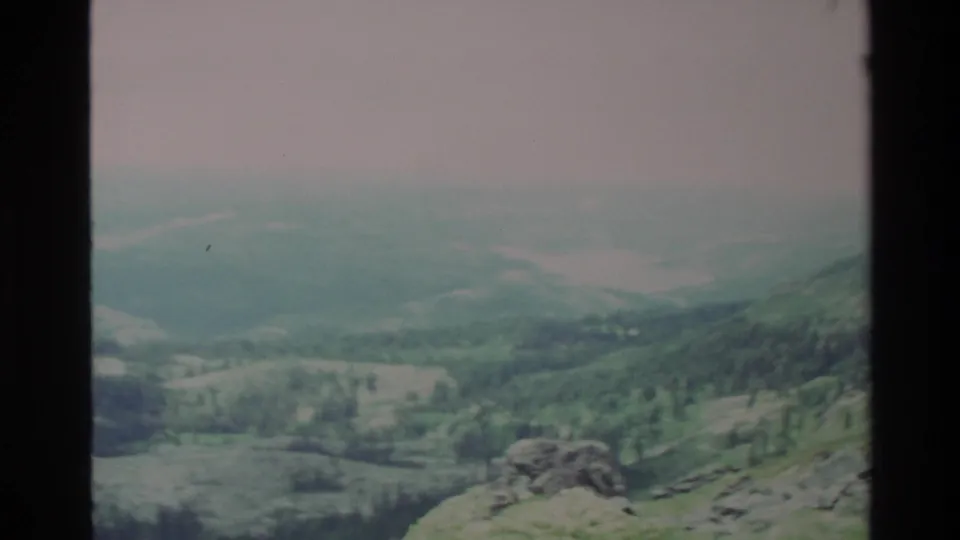 Mid-pan frame from apparent 1980s 8mm footage of Big Sur area; rocky outcrop and dense conifers in mid-ground, pale hazy sky above.