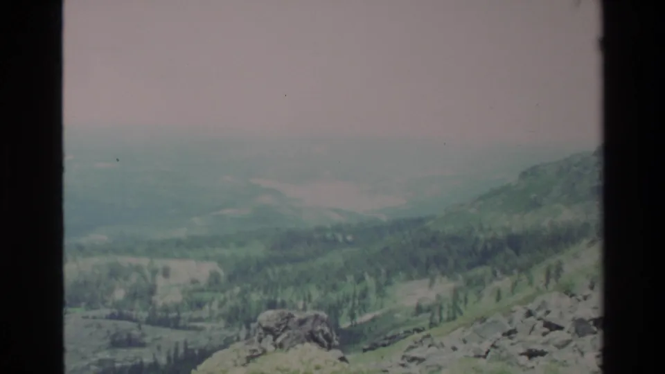 Broader valley view in apparent 1980s Big Sur 8mm home movie; large rocky foreground boulders, forested slopes receding into hazy distance.