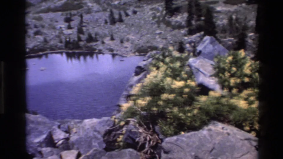 Small alpine pond surrounded by granite boulders and yellow wildflowers, from 1980s 8mm footage attributed to Big Sur, California.