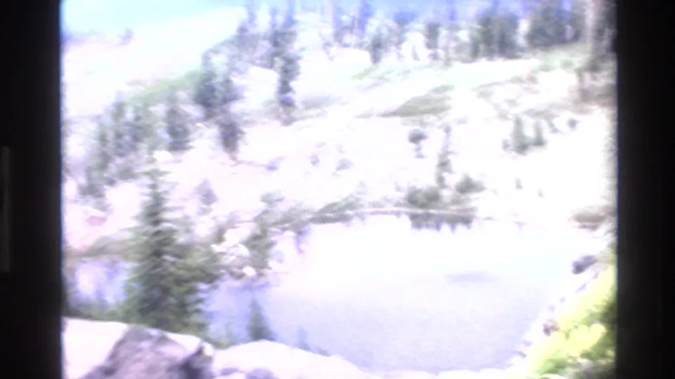 Bright wide-angle view of rocky mountain landscape with sparse conifers and a small alpine pond, from 1980s 8mm footage attributed to California.