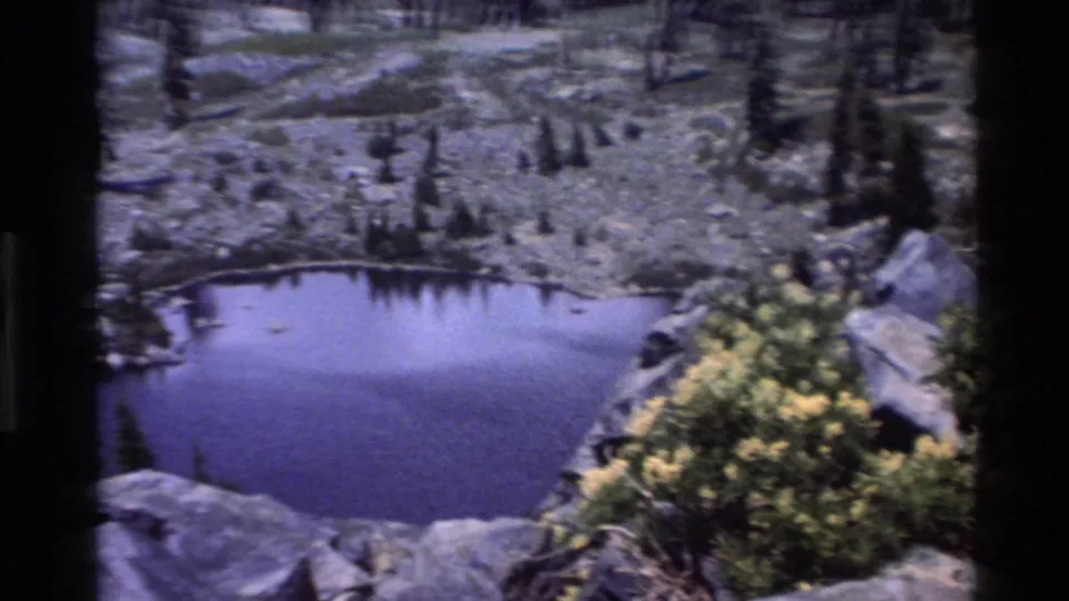 Still alpine pond ringed by granite rock and yellow wildflowers, from 1980s 8mm home movie footage attributed to Big Sur, California.