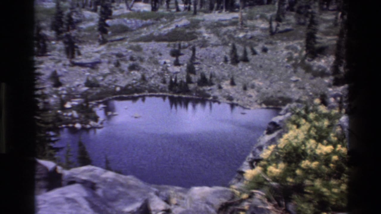 1980s Super 8 shot of a small alpine tarn amid rocky slopes and conifers with yellow wildflowers, attributed to Big Sur, California.