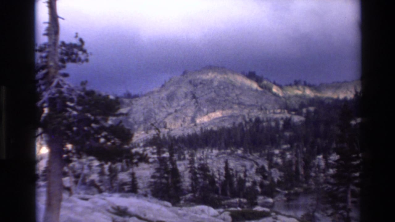 Super 8 home movie frame circa 1980 showing a granite mountain ridge above evergreen forest with overcast blue-purple sky, California wilderness.