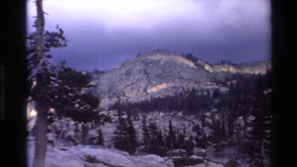Super 8 home movie frame circa 1980 showing a granite mountain ridge above evergreen forest with overcast blue-purple sky, California wilderness.