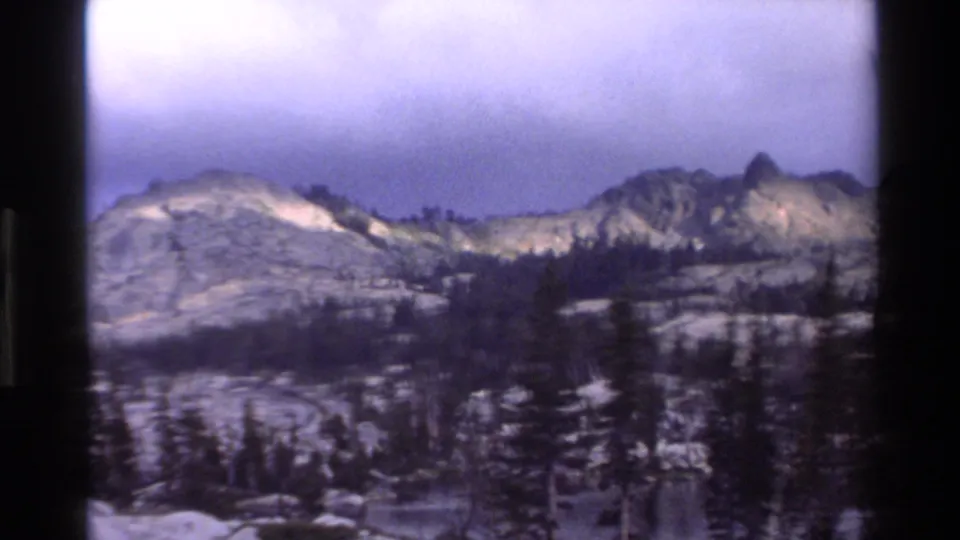 Super 8 frame circa 1980 showing a broader mountain panorama with rugged jagged peaks, evergreen forest, and snow-covered rocky terrain in California wilderness.