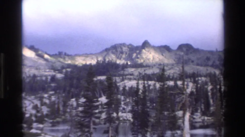 Super 8 frame circa 1980 showing multiple rugged mountain summits revealed by a slow pan, with dense pine forest mid-ground and rocky terrain below.
