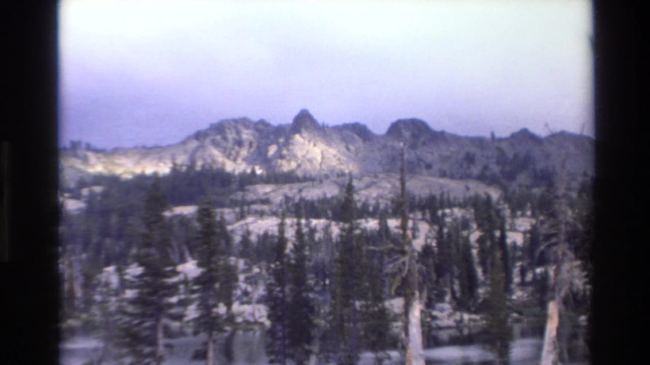 1980s 8mm home movie frame showing jagged mountain peaks, snow-patched slopes, and conifer forest, attributed to the Big Sur region, California.