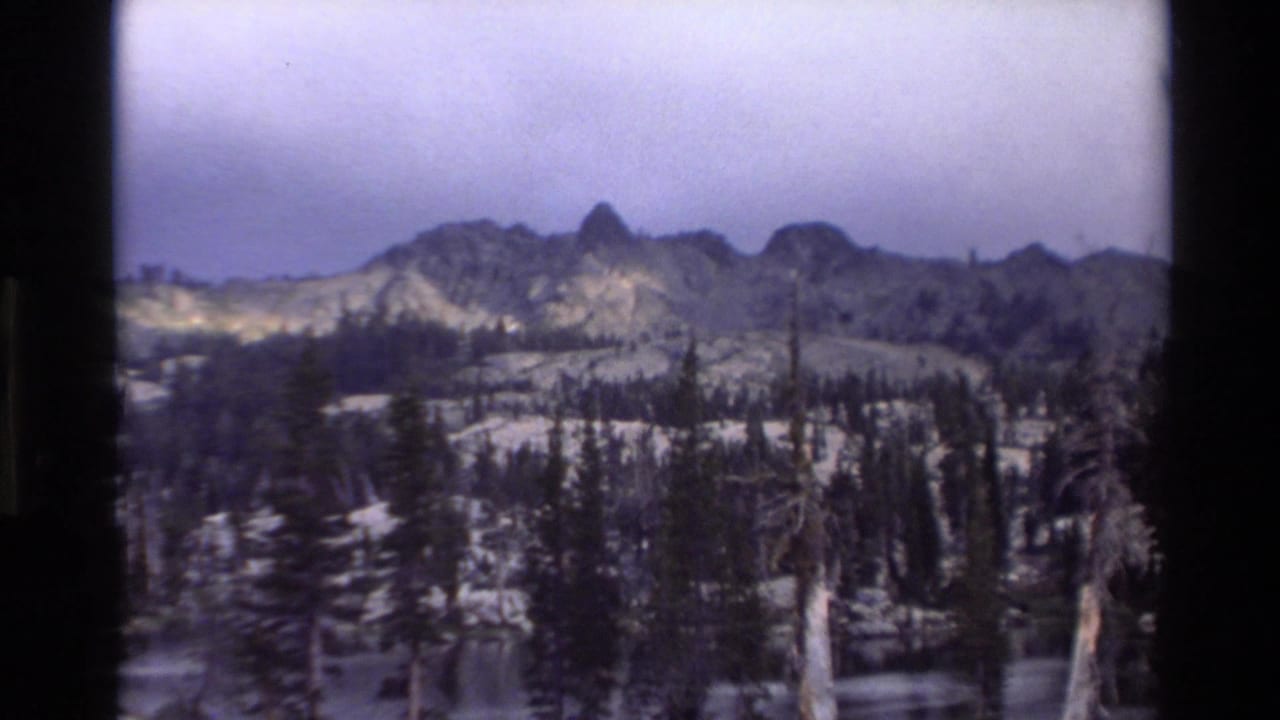 Wide view of rocky peaks and conifer forest with snow patches on the slopes, slight camera settle from previous frame.