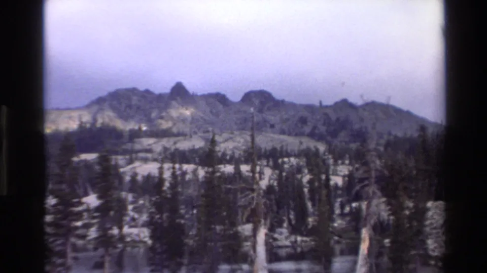1980s 8mm frame with reframed lower angle showing snowy subalpine foreground, dead snag trees, and jagged mountain peaks attributed to California.