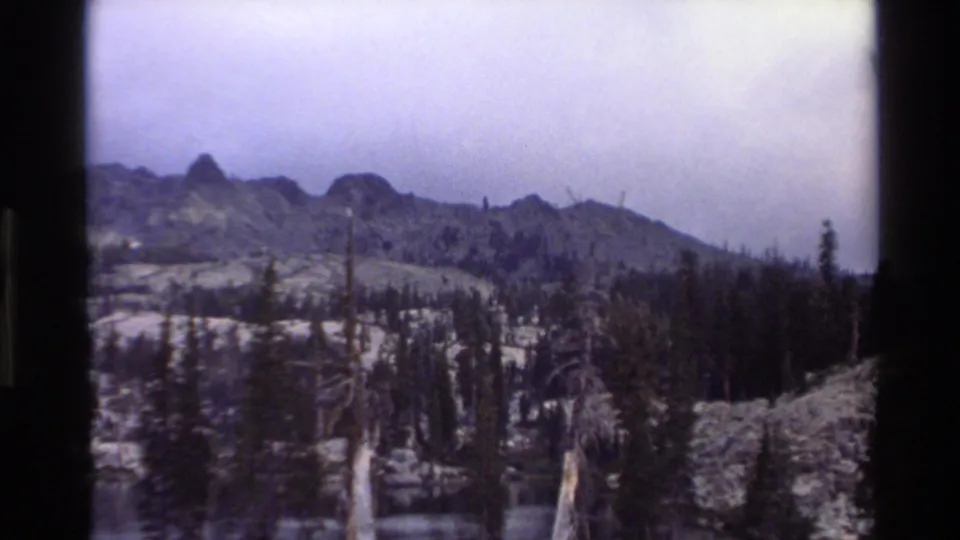 Closing 8mm frame of a California mountain landscape circa 1980, showing jagged peaks, snow-patched subalpine terrain, conifers, and dead standing snag trees.