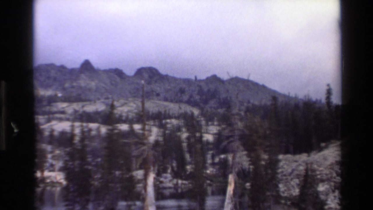 8mm frame showing jagged mountain peaks, snow-covered terrain, and sparse conifers in what appears to be California coastal mountains, circa 1980.