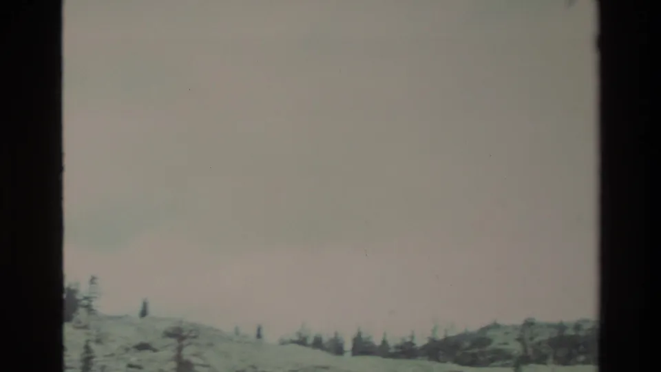 8mm frame from continued upward pan: predominantly pale gray-pink overcast sky with a narrow band of snowy terrain and sparse conifers at bottom, California backcountry circa 1980.