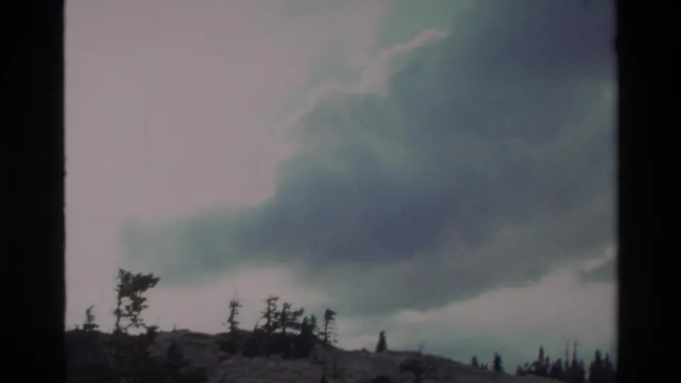 Gnarled conifers silhouetted on a rocky outcrop against a swirling cloud formation; Big Sur highlands, California, circa 1980, 8mm film.