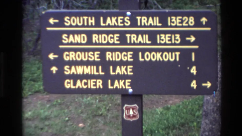 Close 1980 Big Sur 8mm frame of the brown trail sign showing route names, arrows, mileage numbers, grass, trees, and the Forest Service shield.