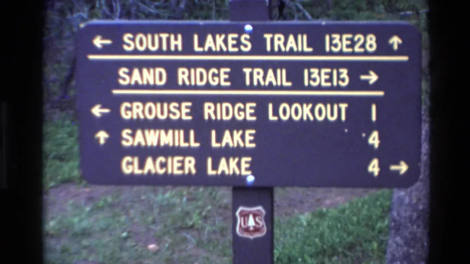 Static 1980 Big Sur home-movie frame of the trail sign with destinations and arrows, set against grass and trees with a U.S. Forest Service shield below.