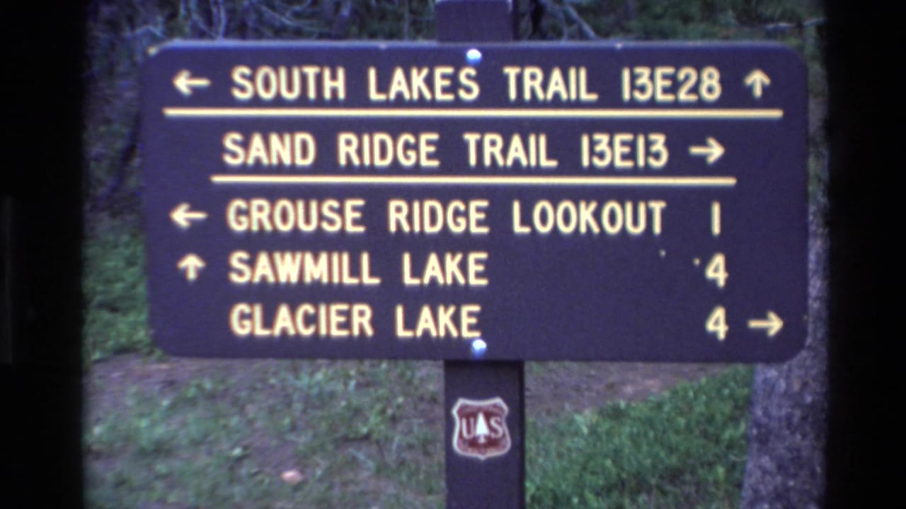 1980 Big Sur home-movie frame showing a dark trail sign listing South Lakes Trail, Sand Ridge Trail, Grouse Ridge Lookout, Sawmill Lake, and Glacier Lake.