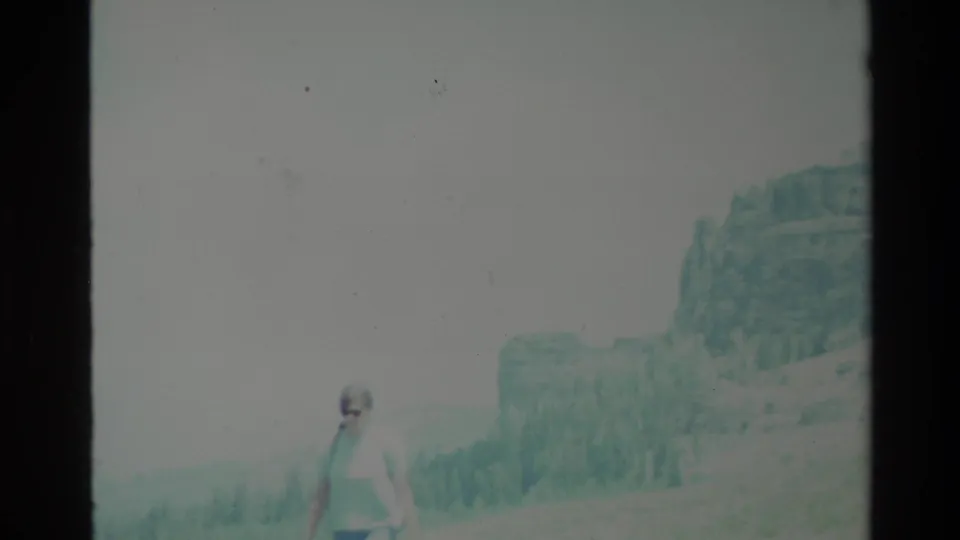 Wide 1980 Big Sur home-movie frame showing a small hiker on a bright hillside with rugged mountain formations in the distance.