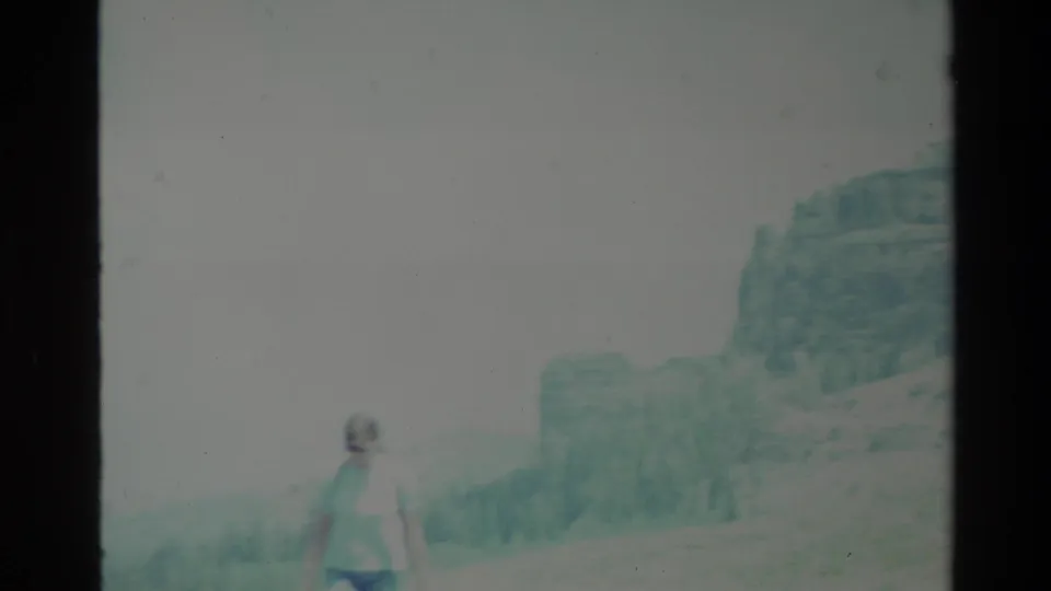 1980 Big Sur home-movie frame showing a woman at left before rugged hills and a washed-out sky.