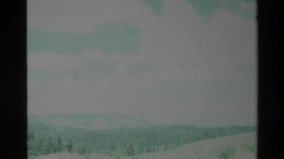 1980 Big Sur 8mm home-movie frame showing distant forested ridges beneath a pale blue sky with soft clouds and dark film borders.
