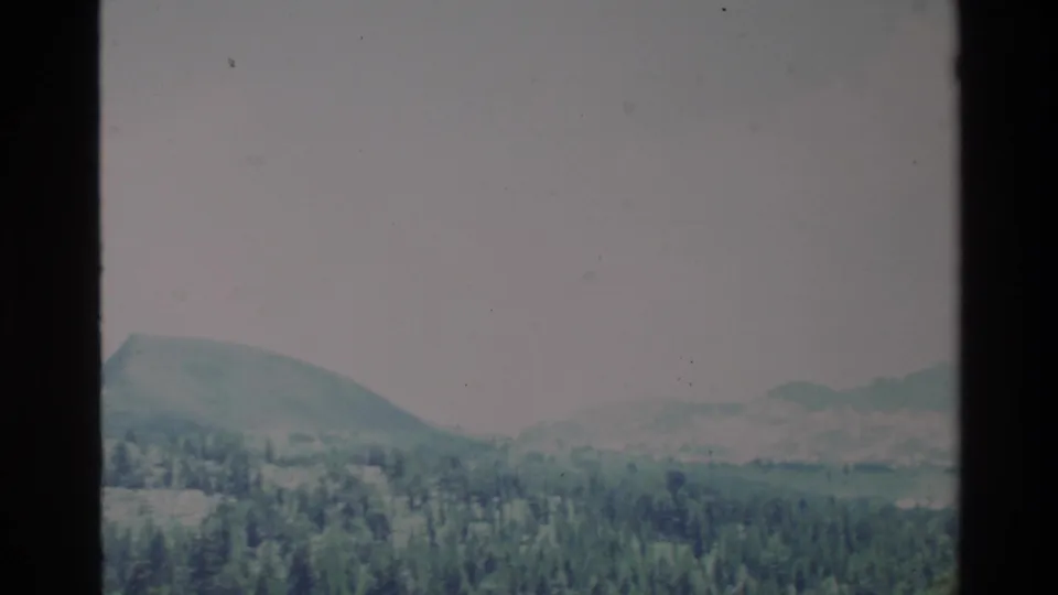 1980 Big Sur 8mm frame with dense forest, rolling hills, and hazy ridgelines under a washed pale sky.