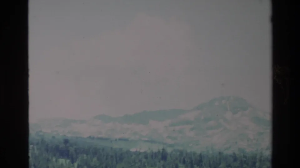 1980 Big Sur home-movie frame with a rugged mountain ridge, layered hills, and dense pine forest below.