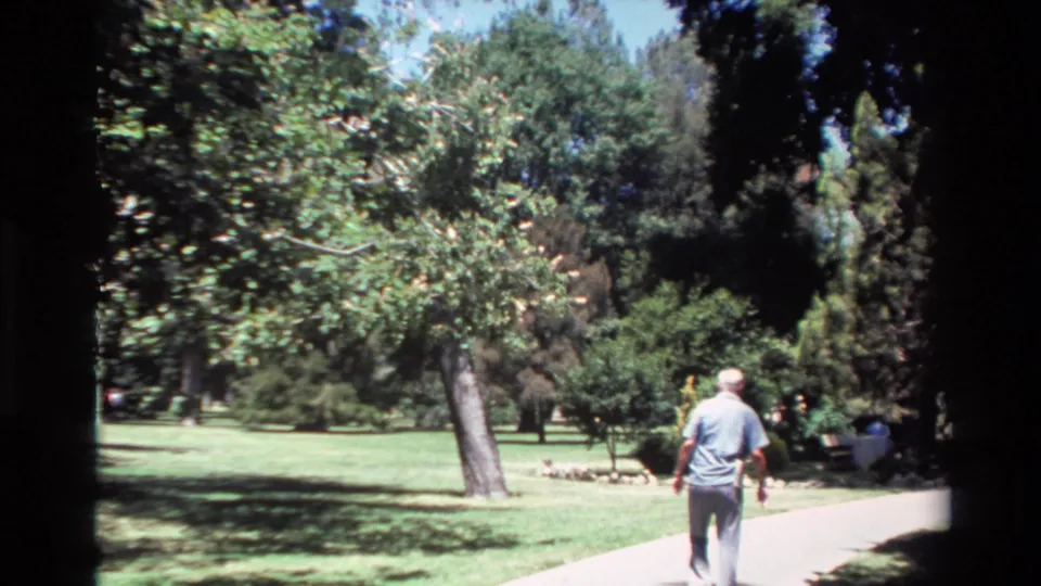 The man continues away from the camera on a paved path bordered by lawn and trees in the 1980 Big Sur home-movie clip.