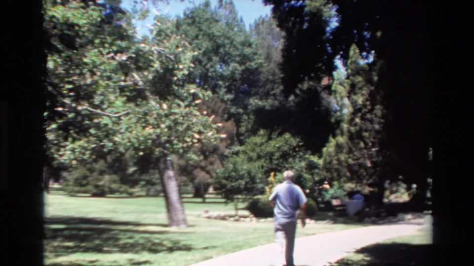 In Big Sur, California home-movie footage, an older white-haired man walks away along a paved path bordered by grass and trees.