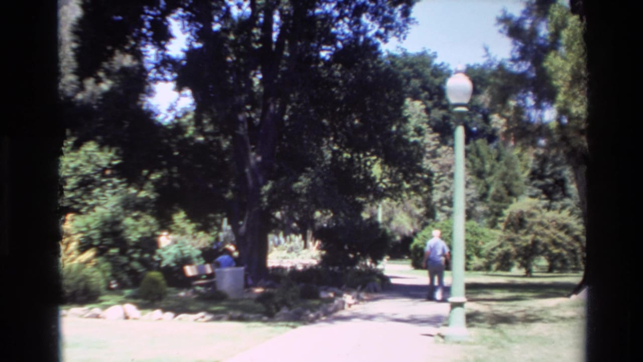 1980 Big Sur home-movie frame showing a person walking along a park path near a green lamppost, with another person seated on a shaded bench.