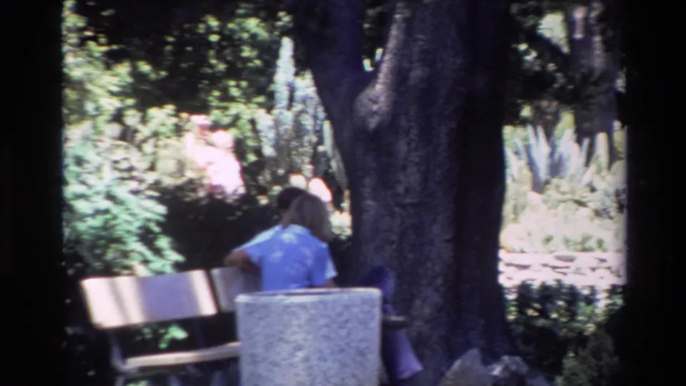 Handheld 1980 Big Sur home-movie frame showing a person on a bench partly obscured by trees in a planted outdoor area.