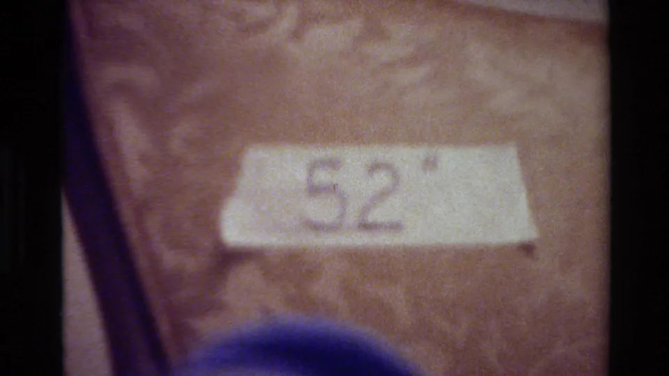 A close 8mm frame shows the handwritten number 52 on white tape, with a blue cylindrical form at the bottom edge and a textured brown background.