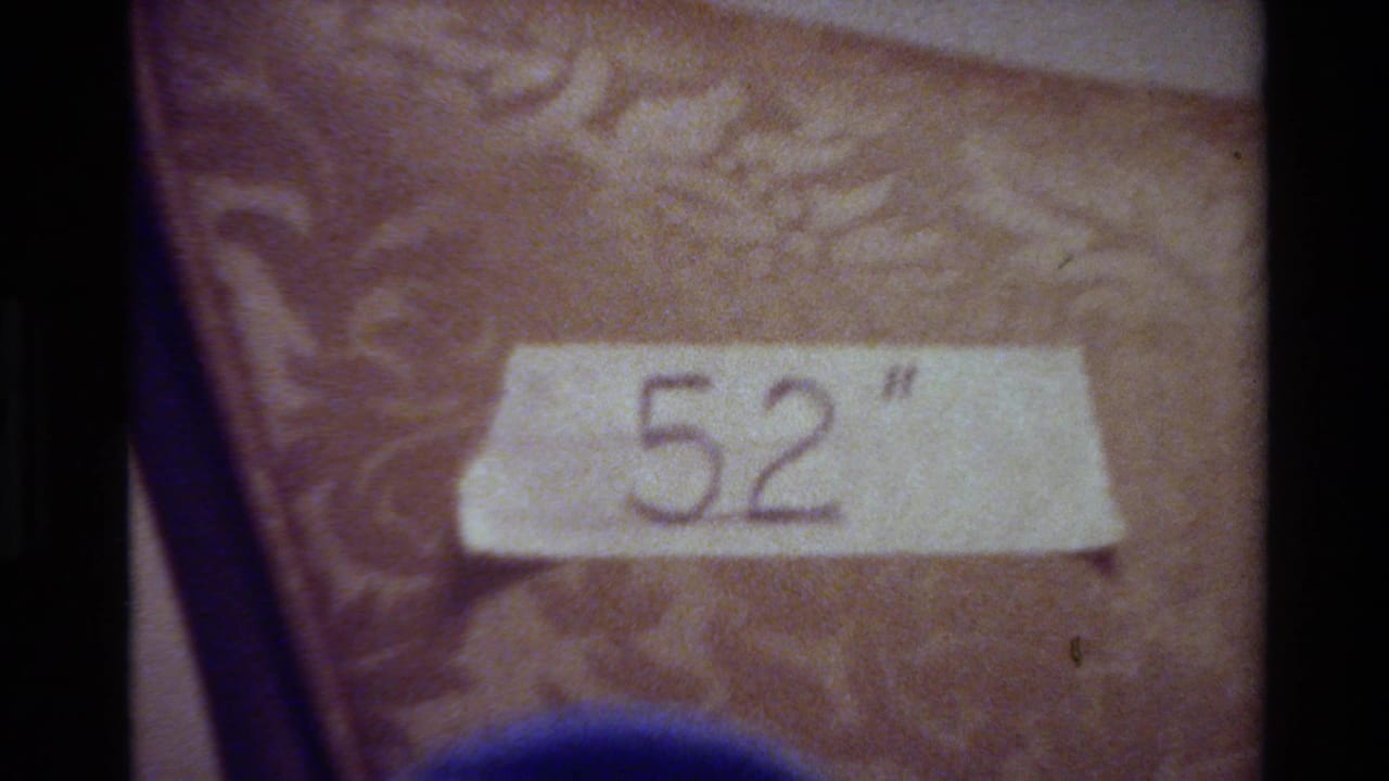 1980 Big Sur home-movie frame showing masking tape marked 52 inches on a patterned chair, with soft focus and film borders.