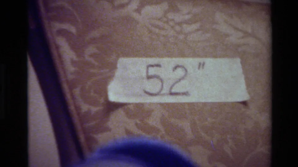 8mm home-movie still of masking tape labeled 52 inches on a patterned chair, partly obscured below by a blue blurred object.