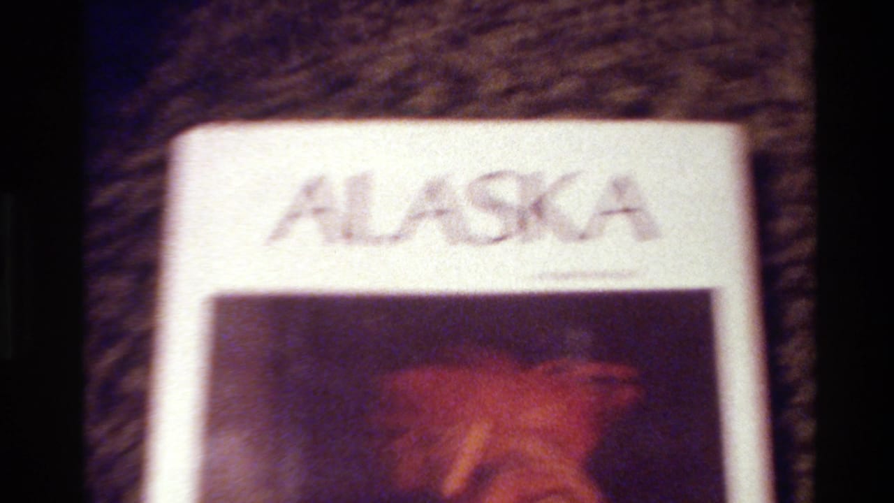 1980 8mm frame from Big Sur showing a soft, partial view of an ALASKA slide or page with a reddish carved object below the title.