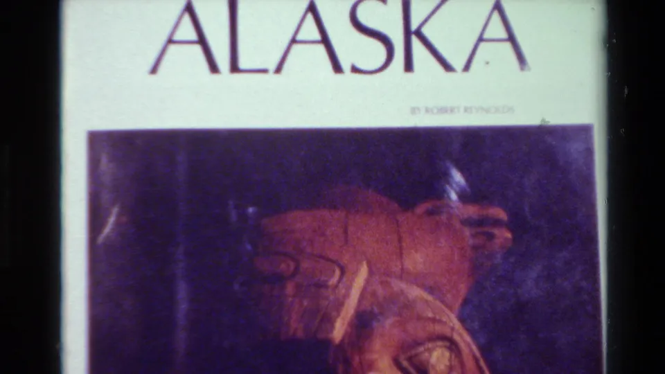 1980 8mm frame from Big Sur showing the ALASKA title, a byline, and a reddish carved object in a projected or filmed slide.
