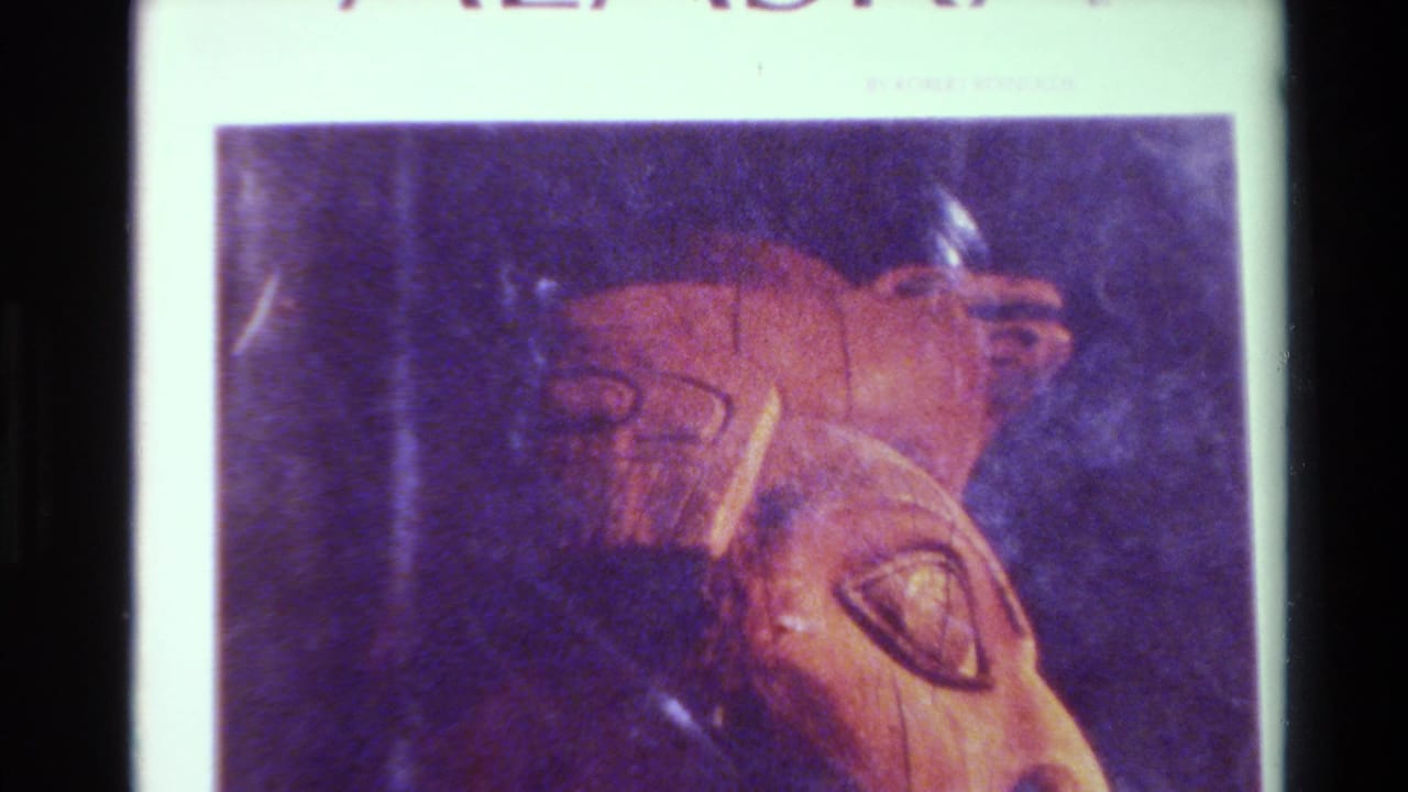 1980 Big Sur home-movie frame showing a close view of a brown carved face on a printed or displayed page with a dark purple background.